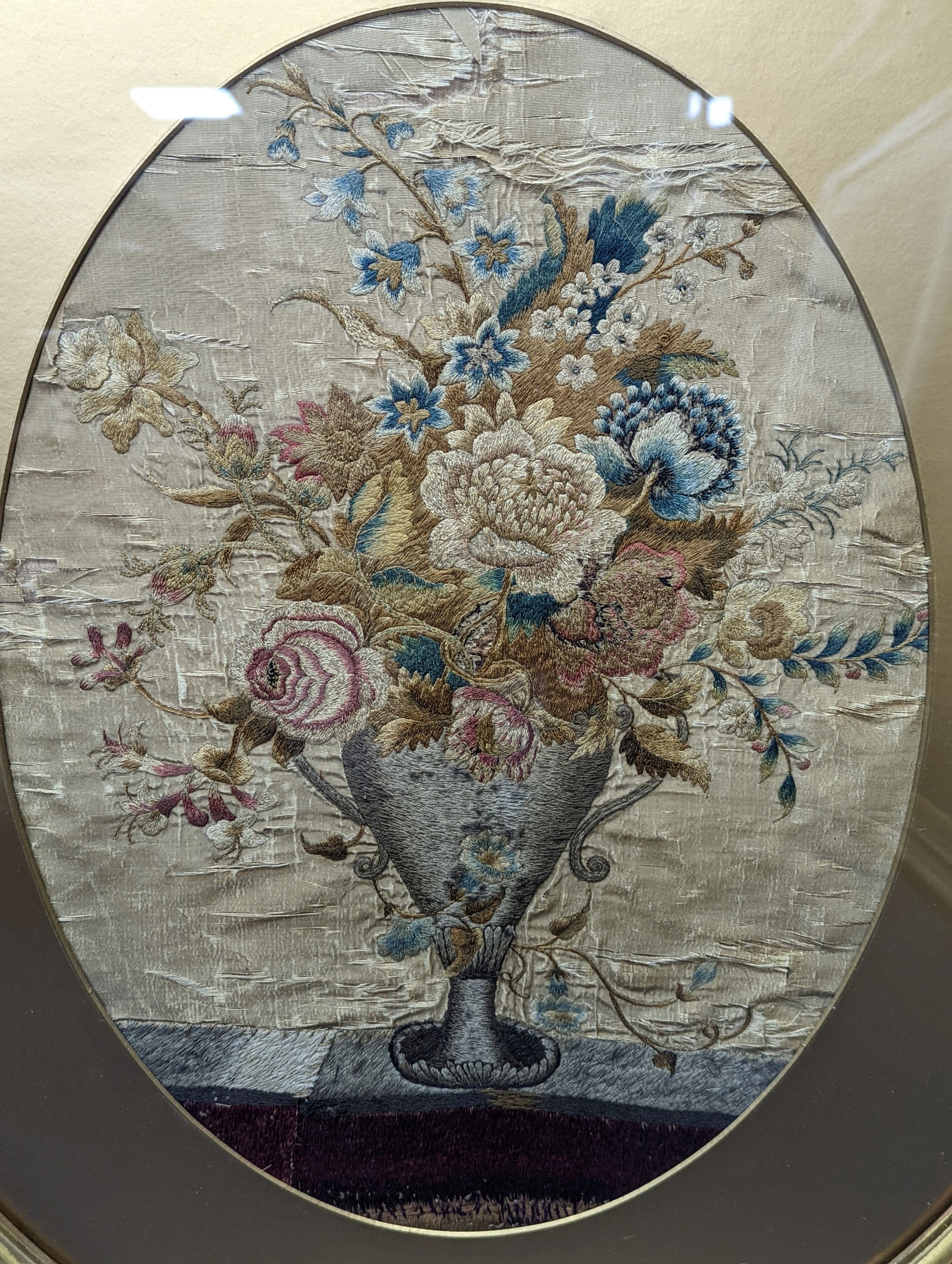 A Georgian oval silk embroidery panel depicting a two handled classical urn containing mixed flowers 46cm x 34cm, glazed gilt frame
